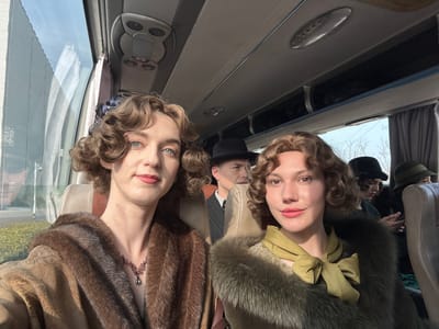 The scene depicts the interior of a bus, likely in motion, within the Huangdao District, China. Bright daylight illuminates the setting through large windows on the left and right. Six individuals are visible, dressed in clothing reminiscent of an early to mid-20th-century period.
In the immediate foreground, two individuals are positioned, looking directly at the camera. The person on the left has light skin, blue eyes, and medium-length curly brown hair. They wear a dark brown fur stole over a patterned garment and a necklace with a dark pendant. The person on the right has light skin, dark eyes, and medium-length wavy brown hair. They wear a large olive green fur stole over a lighter patterned garment, featuring a prominent olive green bow at the neckline. Both individuals are seated in light-colored bus seats.
Behind the person on the left, the upper body of another individual is visible, wearing a dark fedora-style hat and a pinstriped suit jacket, looking forward. Further back, behind the person on the right, two additional individuals are partially visible. One wears a dark cloche hat and appears to be looking down, with hands clasped. The other, further back, wears a dark green cloche hat and looks forward. A sixth individual is faintly visible in the far background, also wearing a hat.
Overhead compartments run along the bus ceiling. Through the windows, reflections of the bus interior and occupants are visible. The exterior environment includes what appears to be a large, light-colored building with reflective surfaces and bare tree branches.