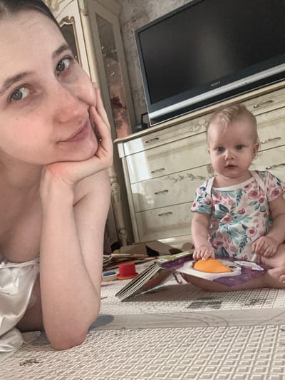 A woman lies on a patterned rug with her hands supporting her chin, taking a selfie. A baby in a floral outfit sits beside her, holding a board book with a cartoon bird on the cover. A TV is mounted on a wall with decorative drawers. The location is inside an unidentified residence, and there's no information indicating it's in Pokrov, Russia.