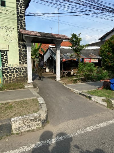 A man in a green shirt and black backpack walks down a paved walkway. He is walking under a concrete archway with white pillars. The archway is attached to a building with a green stucco exterior and a black rock wall. There is a building on the right with a sign that reads "WARUNG NASI" which is located in the city of Purwakarta, Indonesia at -6.556, 107.436. A second man in a blue shirt is sitting on the ground, facing right, with his legs outstretched. The walkway is bordered by grass, shrubs, and a gray sidewalk on the left, and a gray sidewalk and a white painted curb on the right. The sky is a blue, with white clouds, and power lines are visible across the sky.