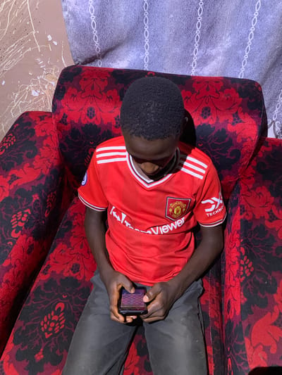A young Black male sits on a red and black patterned armchair in a room. He is wearing a red Manchester United jersey with white stripes on the sleeves and is looking down at a mobile phone he holds in his hands. The armchair has a high back, and the surrounding wall is a light brown color with white paint splatters and some damage. A gray curtain with a white patterned design hangs on the wall behind the armchair. The room appears to be in a residential setting, likely within Barakin Sabawa, Nigeria.
