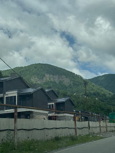 The image captures a view of a construction site with modern houses under construction in the foreground, set against a backdrop of lush green mountains and a cloudy sky. The houses are dark in color with metallic roofs and feature balconies. A concrete wall with rusted metal railings runs along the front of the property, with exposed construction materials and equipment visible behind it, including a yellow excavator. In the distance, the mountains are covered with dense vegetation, and a few small structures are visible on the slopes. The scene appears to be a residential development in a mountainous area. The location is identified as Plastunka, Russia.