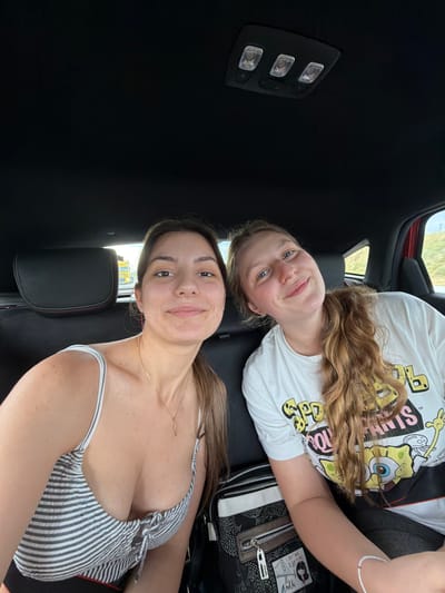 Two young women are in the back seat of a car. The woman on top wears a white SpongeBob t-shirt and has wavy blonde hair. The woman below wears a striped top and a gold necklace. The car's interior is black, and the ceiling light is visible. It is not possible to determine the location of the image is Chokoba, Bulgaria from the image.
