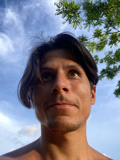A close-up, low-angle shot captures a young man looking upwards against a bright blue sky dotted with scattered white clouds. The man has dark, slightly messy hair that falls across his forehead and temples. His skin appears tanned, and he has a light stubble on his chin and upper lip. The sunlight casts a warm glow on his face, highlighting textures and contours. In the upper right portion of the frame, green leaves and branches of a tree are visible, suggesting an outdoor setting. The image is framed from the chest up, with the sky occupying a significant portion of the background. The scene is devoid of any urban elements or specific landmarks that would indicate a location within Los Velásquez, Venezuela.