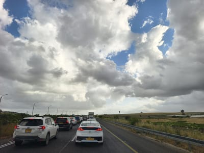 The image shows a traffic jam on a highway in Alona Regional Council, Israel. Several cars are visible, including a white Subaru Forester with license plate 476-12-302 and a white Hyundai Ioniq with license plate 175-28-501. The sky is partly cloudy, with patches of blue visible. The highway is bordered by a metal guardrail and a field with sparse vegetation. Overhead power lines and light posts run along the side of the highway. In the distance, the landscape appears to be agricultural.