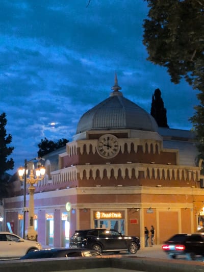 The image shows a building with a dome and a clock, lit by streetlights and the moon. The building appears to be a bank, with a sign that reads "Ziraat Bank". There are several cars parked in front of the building, and two people are standing on the sidewalk. The sky is a deep blue, with some clouds. The location is the city of Ganja, Azerbaijan.