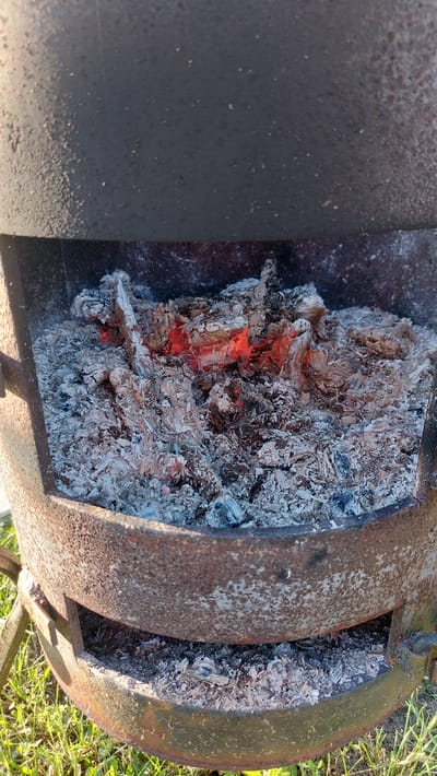 This image shows the interior of a metal brazier containing embers and ash. The brazier is dark gray and appears to be made of thick metal, showing signs of rust and wear, particularly around the rim. Inside, remnants of burnt wood are visible, with glowing red embers interspersed with light gray ash and darker charred pieces. The brazier is positioned outdoors, with green grass and some visible dirt on the ground beneath it. The lighting suggests it is daytime, with sunlight casting shadows. The scene is located in the city of Masty, Belarus.