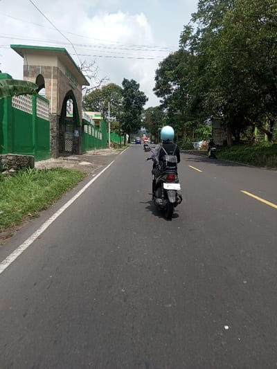 A daytime street scene in Khajuripada, India, depicts an asphalt road extending into the distance under a partly cloudy sky. In the foreground, a person on a dark-colored scooter, wearing a light blue helmet and a black backpack over a dark jacket, proceeds away from the viewer in the right lane. The scooter has a white license plate with black characters. A solid white line marks the right edge of the road, transitioning to a double broken yellow line further down.
To the left, a building with vibrant green walls and a white patterned lattice top runs parallel to the road. An archway entrance, constructed from tan and brown stone-like material and topped with a light green roof, is visible in the midground, featuring indistinct signage. Green grass and sparse vegetation line the roadside adjacent to this structure.
To the right, a dense canopy of green trees borders the road. Further down this side, another individual is visible on a parked or slowly moving motorcycle. In the far background, vehicular traffic is visible on the road, and distant, hazy hills are discernible against the sky.