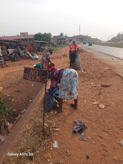 The image depicts a roadside scene in Awkuzu, Nigeria, characterized by market activity and ground-level commerce. In the foreground, a crudely constructed "NO PARKING" sign on a metal pole is prominent on the left. The ground is reddish-brown dirt, dry and sparsely vegetated, with scattered litter including fabric scraps, plastic, and paper.
Three women are central to the scene's midground. One woman, in the immediate foreground, wears a patterned long-sleeved top and a light blue and brown patterned skirt, bending low to sort or pack items into a dark grey sack. Her feet are visible in dark flip-flops. Slightly behind her, another woman in a floral dress is similarly crouched, near a green basin or container, engaged in a packing activity. Further right, a third woman stands upright, her back to the viewer, wearing a red jersey with the number "11" and dark bottoms, positioned next to a large, light-colored sack filled with contents.
To the left, a series of open-air market stalls with wooden frames and corrugated metal roofs are visible, surrounded by numerous large sacks, suggesting a market area. A sign, partially obscured, reads "Paddy Market." Multiple individuals are seated or standing within this market section.
To the right, a two-lane paved road extends into the distance. Vehicles, including a large green truck and two motorized tricycles ("Keke Napep"), are observed moving away from the camera on the road. The shoulder of the road is unpaved and consists of reddish-brown dirt. In the distant background, a tall metal communication tower stands against a hazy, pale sky, with scattered green foliage visible across the landscape. The overall lighting suggests an outdoor environment, possibly under a bright or slightly overcast sky.
