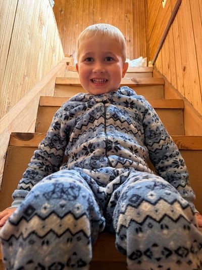 A young boy with blond hair sits on a wooden staircase, wearing a blue and white patterned onesie. He is smiling and looking directly at the camera. The staircase is made of unfinished wood and is located indoors, likely in a rustic dwelling, as indicated by the wood-paneled walls. The environment suggests a residential setting in the village of Papiarnianski Sel'ski Savet, Belarus. The lighting is warm and natural, casting soft shadows.