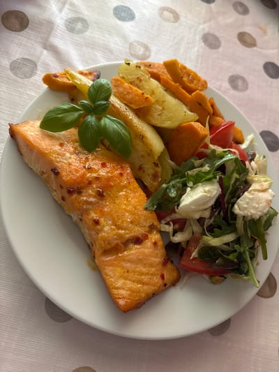 A white plate holds a meal of salmon, roasted vegetables, and a salad, placed on a tablecloth with a polka dot pattern. The salmon fillet, seasoned with red flecks and herbs, is on the left side of the plate. To the right of the salmon are roasted potato wedges and sweet potato wedges. A fresh salad composed of mixed greens, tomatoes, and mozzarella balls is situated on the far right. A sprig of fresh basil rests atop the roasted vegetables, serving as a garnish. The setting is indoors, on a table covered with a light-colored tablecloth adorned with large, grey polka dots. The lighting suggests it is daytime. The meal is likely prepared and photographed in a residential kitchen or dining area in Wörth am Main, Germany.