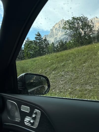 The image is taken from inside a vehicle, looking out the window towards a mountainous landscape. The foreground shows the interior of a car door, including a speaker grille and seat adjustment controls. The window is speckled with water droplets, suggesting it might be raining or has recently rained.
Outside the window, a grassy slope leads up to a backdrop of rugged, rocky mountains. Several trees, mostly coniferous, are scattered across the slope and at the base of the mountains. The sky above is partly cloudy. A side-view mirror is visible, reflecting a sliver of the road and landscape.
The scene is consistent with a scenic drive in a mountainous region. The style of the car interior suggests a modern luxury vehicle. Given the context provided about San Vito di Cadore, Italy, this view likely depicts the Dolomites mountain range, a popular tourist destination known for its dramatic peaks and alpine scenery. The image captures a moment of travel, observing the natural beauty of the Italian Alps through the filtered lens of a car window.