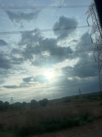 The image depicts a landscape with power lines crisscrossing the frame. In the foreground, dry grass and some green vegetation cover the ground, suggesting an agricultural or rural setting. In the mid-ground, there are trees and rolling hills. Several power transmission towers are visible in the distance, including one prominent tower on the right side of the image. The sky is cloudy, with the sun partially obscured by clouds, casting a diffused light. The scene appears to be located in or near Sugali Tanda, India, as indicated by the presence of typical rural Indian landscape elements and infrastructure. There are no people visible in the image.