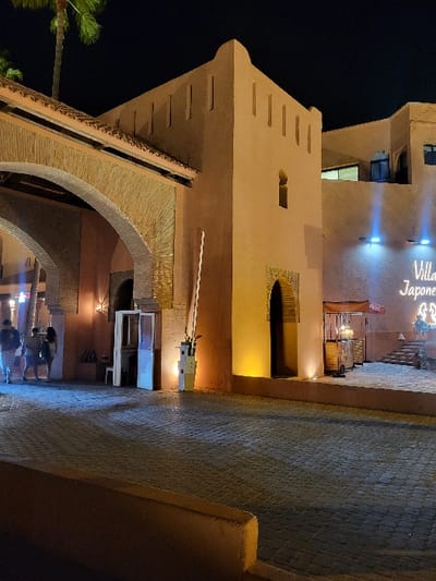This night-time image depicts the illuminated entrance to a large, earth-toned building complex in Bouznika, Morocco. The structure displays traditional Moroccan architecture, featuring a prominent brick archway on the left and a central tower-like section with crenellated parapets and small vertical apertures. A partial neon sign on the right wall reads "Japone." The foreground is paved with dark interlocking bricks. A raised white barrier arm is present in the open position near a grey control box. Four blurred human figures are visible beneath the archway on the left, their activities indistinct. The complex is lit by external warm yellow uplights and blue downlights, casting shadows. A tall palm tree is partially visible on the far left. A small white booth is located left of the barrier, and a covered stand or kiosk sits on a raised platform to the right, accessible by stairs.