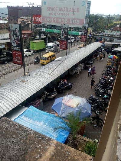 An elevated view shows a busy street and marketplace in Cipanas, Indonesia. A yellow van drives along the road, passing by stalls covered by a long, metal awning. People stroll along the market, while motorcycles fill parking spaces. Billboards advertise "SPACE AVAILABLE" with phone numbers and feature the phrase "Ayo Belanja ke PASAR RAKYAT CIPANAS" which translates to "Let's Shop at CIPANAS PEOPLE'S MARKET." An overhead pedestrian walkway crosses the street.