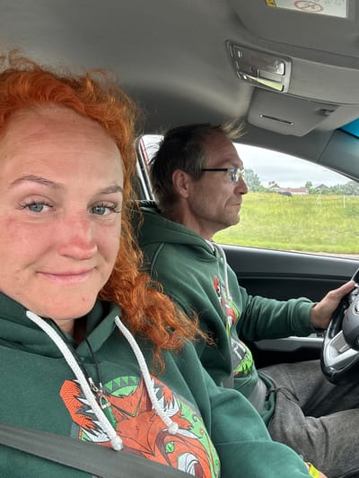 An outdoor, eye-level, medium shot captures two individuals inside a car, possibly traveling on a road in the vicinity of Alnashi, Russia. The woman in the foreground, positioned on the left, has bright orange, curly hair and is looking directly at the camera with a slight smile. She is wearing a green hooded sweatshirt with a colorful graphic design and a grey seatbelt. Her face shows some freckles and appears to be slightly flushed.
To her right, the man is driving the car and looking forward. He has short, light brown hair, wears glasses, and is also dressed in a green hooded sweatshirt with a similar graphic. The interior of the car is visible, including the dashboard and a sun visor.
Through the car windows, a rural landscape is visible. The scenery includes a grassy field with scattered trees and some distant buildings, suggesting a countryside setting. The sky is overcast, indicating potentially cloudy weather. A single dark-colored animal, possibly a cow, is grazing in the field in the distance. The overall scene suggests a casual road trip.