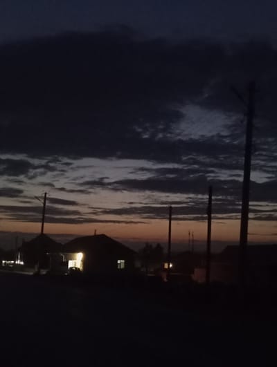 The image depicts a dark, twilight scene in what is identified as the city of Kızıl-Bel, Kyrgyzstan. Silhouetted against a dusky sky are several residential structures, the roofs of which are dimly visible. One house in the center of the frame is illuminated from within, with a single light source casting a yellow glow. A car is parked in front of this illuminated house. Power lines and poles are prominent in the foreground and midground, extending upwards into the cloudy sky. The sky itself is a blend of dark blues and grays, with hints of orange and yellow near the horizon, suggesting either sunrise or sunset. The overall atmosphere is serene and quiet, characteristic of a rural or suburban setting at the end of the day.