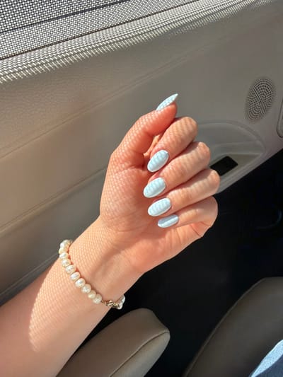 The scene depicts a person's left hand and wrist inside the passenger compartment of a vehicle. The hand is illuminated by direct sunlight filtered through a mesh screen, casting a distinct grid-like shadow pattern across the skin. Five almond-shaped fingernails are visible, each painted light blue with a white wavy or striped pattern. A pearl bracelet, composed of irregular, light-colored pearls secured with a gold-toned clasp, is worn on the left wrist. The immediate environment includes a light-colored (likely beige or off-white) car door panel on the left and upper right, featuring a circular speaker grille with a fine mesh pattern on the upper right section. A beige or tan upholstered car seat component, possibly a headrest or armrest, is visible in the lower mid-right. A dark area in the lower center likely represents the vehicle's floor. A small segment of blue denim fabric is visible in the lower right corner. No specific visual cues within the image allow for the identification of a precise location within Veracruz City, Mexico.