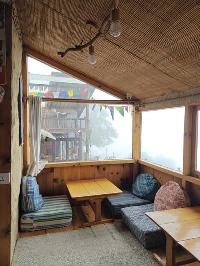 This image depicts a cozy, rustic seating area within a building in Prini, India. The space features a low table made of natural wood, surrounded by floor cushions and seating mats. The walls and ceiling are constructed from wood and bamboo, creating a warm and organic ambiance. Prayer flags are visible strung across the upper part of the window, suggesting a cultural or spiritual context. The large windows offer a view of a misty, overcast landscape, with hints of buildings and trees visible through the fog. The overall impression is one of a serene and comfortable retreat, possibly a cafe or guesthouse. There are no people present in the image.