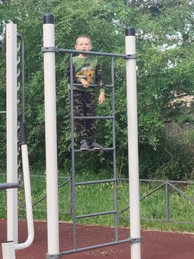 A young boy stands on a ladder attached to outdoor exercise equipment. He is wearing a long-sleeved green shirt with a graphic of a yellow vehicle on it, and camouflage pants. The boy is looking towards the camera with a neutral expression. The exercise equipment is silver metal with white poles. Behind the equipment, there is lush green foliage. The ground in front of the equipment is covered in a reddish-brown material. The scene appears to be in a park or playground setting. This image was taken in Askiz, Russia.