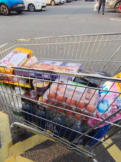 This image captures a shopping trolley filled with groceries, placed on a grey asphalt surface marked with a yellow line, likely in a supermarket parking lot. The trolley contains a variety of items, including a pizza box, boxes of Balconi Trancetto Cacao biscuits, a carton of eggs, bottles of liquid, and other packaged foods. A person's legs and lower torso, wearing dark trousers and shoes, are visible in the background, carrying a white plastic bag. The sunlight casts shadows, suggesting it is daytime. The background shows parked cars, including a blue SUV and a white car, with license plates visible. The overall scene depicts a common consumer activity of grocery shopping. Visible text includes brand names like "Balconi," "Farm Foods," and "Gill Beck." The overall impression is that of a mundane, everyday errand.