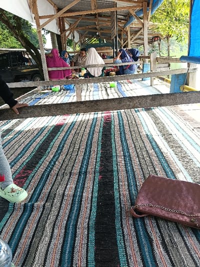 The image depicts an outdoor, semi-enclosed, elevated structure, likely a restaurant or cafe, in Gembong, Indonesia. The structure features a corrugated metal roof partially covered by white and blue tarpaulins, supported by rough wooden beams and posts. The floor is covered with a striped mat, prominent in the foreground, displaying vertical bands of black, blue, teal, white, grey, brown, and hints of red.
Approximately 6-8 individuals are seated on mats in the midground. On the left, two women in hijabs are visible; one wears a magenta garment and hijab, facing right, while another wears a blue patterned hijab. In the central area, a man with glasses and a striped shirt faces a woman in a light-colored hijab. Further right, at least two additional women are seated, one in a dark blue hijab. Small green plastic bowls are placed on the mats near these groups.
In the immediate foreground, the left hand and forearm of an unseen person rest on a wooden railing. This person's left foot, wearing a light green sandal, is also visible. A dark pink, textured clutch bag with a silver chain strap lies on the striped mat in the lower right foreground.
In the background to the left, a black pickup truck is partially visible. Beyond the structure, lush green trees and foliage are present. A body of water is discernible on the far right, beyond a blue tarpaulin barrier and wooden railing, indicating a lakeside or waterfront setting. The scene conveys a casual gathering or meal.