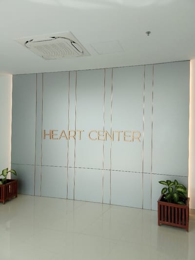 The image shows the interior of a building, likely a hospital or clinic. It is located in the city of Kecamatan Margorejo, Indonesia. The scene is dominated by a large, light-gray tiled wall with thin vertical gold lines spaced evenly across its surface. In the center of the wall is a gold-colored sign that reads "HEART CENTER". The wall is divided into twelve tiles arranged in a 3x4 grid. The floor is a light beige tile, matching the overall clean and modern aesthetic of the space. Two potted plants, one on the left and one on the right, stand near the wall. There are no people in the image. The ceiling is white and features a rectangular air vent. There is a single light fixture in the center of the ceiling. The interior lighting is bright and uniform, creating a clean and sterile atmosphere. The scene is typical of a medical facility, suggesting a focus on cleanliness and organization.