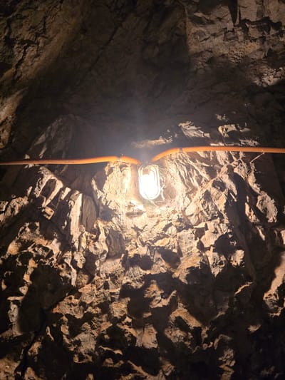 The image shows the rough, textured rock ceiling of a cave or tunnel. A single light fixture, a bulb in a protective cage, hangs from the ceiling and illuminates the scene with a warm, yellow light. Orange corrugated tubing runs horizontally across the ceiling, likely carrying electrical wires. The rock formations are uneven and jagged, with deep shadows creating a dramatic contrast with the lit areas. The environment appears damp and enclosed, typical of an underground space. The scene is devoid of people. The specific location in Perazića Do, Montenegro, suggests this might be a natural cave or an old mining tunnel, possibly in a coastal or mountainous region given the geography of Montenegro.