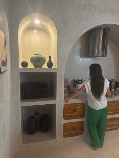 An interior scene depicts a modern kitchen or kitchenette area located in El Nido, Philippines. The walls and integrated shelving are constructed from a light grey, textured material resembling polished concrete, featuring prominent arched recesses.
One individual, a person with long, dark, wet hair, is visible from behind, positioned at a light-colored countertop. They wear a white short-sleeved t-shirt and loose-fitting green trousers, and appear to be barefoot. Both hands rest on the countertop, suggesting a pause or preparation.
To the left, a built-in shelving unit with three levels is present. The top level, an arched niche illuminated by a recessed light, displays three decorative vases: a large, textured, light teal vase centrally flanked by two smaller, dark, simple vases. The middle shelf holds a black LG microwave oven. The bottom shelf contains two dark, distinct decorative objects, one teardrop-shaped and one cylindrical. A partially visible framed artwork hangs on the far left wall.
Above the countertop, a stainless steel cylindrical range hood branded "TEKA" is mounted within an arched recess. The countertop itself holds various kitchen items, including a light-colored blender, a white utensil holder with kitchen tools, and a small bottle among other indistinct objects. Below the countertop, three wooden cabinet doors with rounded corners and circular knobs are visible, suggesting storage. The floor matches the light, textured aesthetic of the walls.