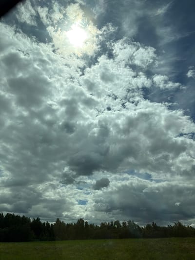 The image shows a cloudy sky with the sun peeking through the clouds. A thin strip of trees and green grass appears on the right side of the image. It is daytime. The location is Юзуфоўскі сельскі Савет, Belarus. There are no people or actions visible in the image.