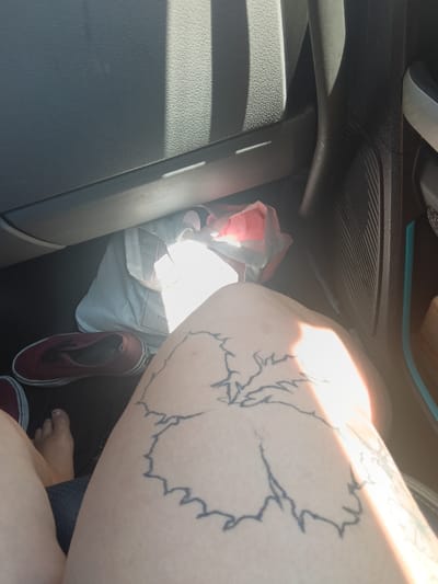 A close-up shot shows the lower part of a person's legs and thighs, adorned with a large, black outline tattoo of a hibiscus flower on the right thigh. The person is wearing dark blue shorts. To the left, a pair of dark red canvas sneakers is visible, with one shoe in focus and the other partially obscured. A white tote bag with a red object inside it is placed on the floor of a vehicle. The scene is illuminated by sunlight streaming through the car window, creating distinct shadows on the dark interior surfaces. The location is the interior of a vehicle, likely parked, in Rock Springs, United States of America.