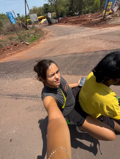 A sunny, outdoor scene features two people on a road, apparently in Marra, India, as indicated by a sign in the background. A person wearing a dark gray t-shirt with a yellow strap across the chest and a gold chain on the arm is holding the camera, capturing a selfie. This person is facing the camera with a neutral expression. Another person wearing a yellow shirt and black shorts is sitting behind them, facing away from the camera. The road is mostly gray and shows signs of wear. The left side of the frame contains vegetation, utility poles, and road signs, while the right side mainly features the road surface.