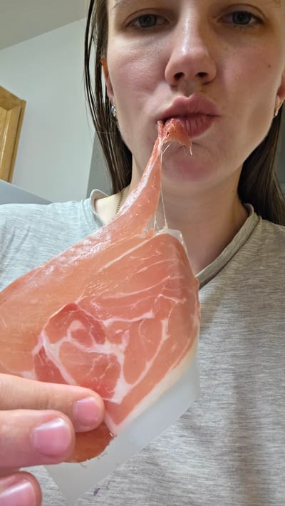 The media file shows a close-up, upside-down perspective of a person. The person is holding a thin slice of prosciutto up to their lips, with a piece of the meat touching their mouth. The prosciutto is pink with white marbling. The person is wearing a gray t-shirt. Their fingernails are visible, appearing short and neat. A delicate gold necklace is visible on their neck. The background appears to be an indoor setting with a light-colored wall and a wooden element near the top of the frame. The lighting suggests it is daytime. The person's facial expression is difficult to discern due to the angle and the prosciutto. There is no visible text in the media file. The image was likely taken in Portland, United States of America.