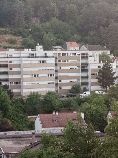 This image captures a residential area in Nancy, France, with a prominent multi-story apartment building as the central focus. The building, featuring a light-colored facade with horizontal bands of beige, is surrounded by lush green trees and foliage. Several cars, including a white van, are parked in front of the building. In the background, a dense forest covers a hillside, and a small, dilapidated stone structure with an open doorway is visible amongst the trees, accessed by a steep, uneven staircase. Smaller residential buildings with red-tiled roofs are situated in the foreground and to the right of the apartment building, some partially obscured by trees. The sky is overcast, suggesting a cloudy day.