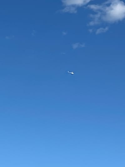 A white drone is captured mid-flight against a clear blue sky with a few white clouds. The drone is positioned in the center-right of the frame, appearing small due to its distance from the camera. The scene is an open sky, with no discernible city structures or land features visible. The location is specified as Слава Снт, Russia.