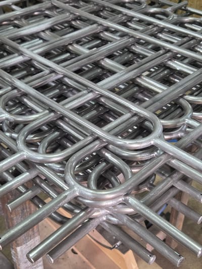 This media file displays a close-up, angled view of a pile of metallic, cylindrical objects. The objects appear to be industrial components or parts, possibly made of steel or aluminum, due to their silvery-gray color and the way light reflects off their surfaces. They are arranged in a somewhat chaotic but interlocking manner, creating a complex visual pattern of lines and curves. Many of the pieces have a U-shaped or looped form, suggesting they might be part of a larger assembly or a structural element. Some of the cylindrical ends are visible, and a few have small holes or indentations. The surface of the objects shows a matte finish with areas of sheen where light directly hits. The background is blurred but suggests an indoor setting with wooden surfaces and a concrete floor, indicative of a workshop, factory, or storage area. There is no clear indication of time of day or weather. The overall impression is one of manufactured goods, possibly awaiting assembly or distribution. No people are visible, and there is no discernible text in the image. The location context provided, Гаранскі сельскі Савет, Belarus, suggests this is likely a scene within that rural administrative area in Belarus.
