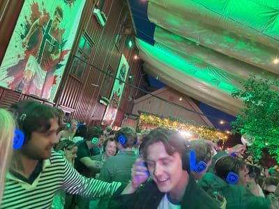 In Tønsberg, Norway, a group of young adults is gathered outdoors in a dimly lit environment, possibly a bar or event space, illuminated by green and yellow lights. Many of them are wearing noise-canceling headphones that feature glowing blue or green logos, suggesting they are participating in a silent disco or a similar audio-focused event. The attendees appear to be engaged and enjoying themselves, with some smiling and looking around. In the background, a large banner featuring a football player in a Norwegian national team jersey is visible on a wooden-paneled building. The scene is set under a large, draped canopy, and trees with foliage are present, indicating an outdoor or semi-outdoor setting. The overall atmosphere is festive and social.