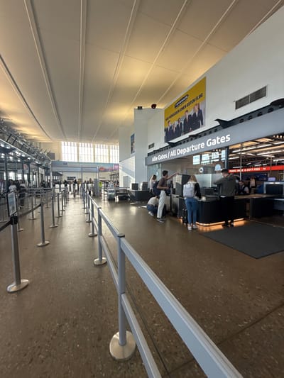 The image shows an interior view of an airport terminal in Künersberg, Germany. The scene depicts a security checkpoint area with queuing barriers and several individuals undergoing screening. To the right, a counter is set up with a scanner, and a person in uniform appears to be assisting passengers. Overhead, a large banner advertises Ryanair with the text "WIR WÜNSCHEN EINEN GUTEN FLUG" (We wish you a good flight). Above the checkpoint, signage reads "Alle Gates / All Departure Gates". The terminal features a high, curved ceiling with exposed lighting and ventilation systems, and the floor is a speckled terrazzo material. Natural light streams in from large windows in the background, illuminating the space. Several people are visible in the queues, some with luggage, and others interacting with security personnel.