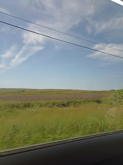 The image captures a rural landscape, likely a field in Craghyboyle, Ireland, seen from a moving vehicle. Two dark power lines bisect the upper portion of the frame, stretching horizontally across a pale blue sky dotted with wispy white clouds. Below the sky, a vast expanse of green fields unfolds, with a darker, possibly plowed or barren strip of land visible in the mid-ground. The foreground is dominated by blurred green vegetation, suggesting the motion of the viewer. A dark, reflective surface, possibly a car's interior or window frame, runs along the bottom edge of the image, further emphasizing the perspective of being inside a vehicle. The scene is devoid of any people or immediate signs of human activity, presenting a serene, natural environment.