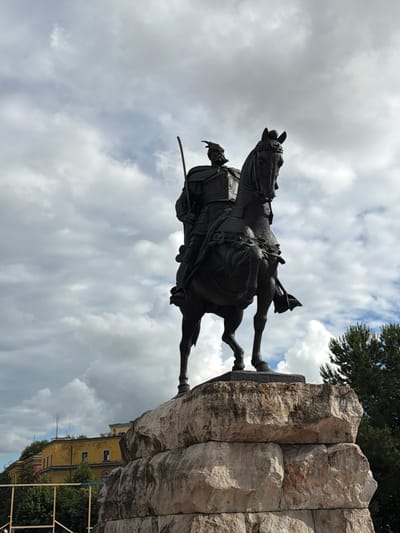 The image, taken from a low angle looking upwards, captures a large, dark equestrian statue against a partly cloudy sky. The statue, likely made of bronze or a similar dark material, depicts a male rider on horseback. The rider appears to be a historical military figure, dressed in what looks like a uniform or armor, and holds a sword or saber at his left side. The horse is sculpted in a dynamic pose, suggesting movement, and is oriented towards the left of the frame.
The monument stands on a robust pedestal constructed from large, light-colored, roughly cut stone blocks. In the upper right background, a dark green coniferous tree is visible, extending into the frame. In the lower right, behind the statue's base, a multi-story yellow building with windows and a flat roof is partially visible. The sky features soft grey clouds with occasional patches of blue, indicating daylight. No discernible text is visible on the statue, pedestal, or in the surrounding scene. The image portrays a prominent historical monument in what appears to be an urban setting, characteristic of places like Tirana, Albania.