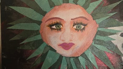 The image displays a close-up, front-facing view of a mixed-media artwork. The central subject is a stylized, light peach-toned face with large green eyes featuring black pupils and prominent dark eyelashes, distinct arched eyebrows, and closed magenta lips. The face exhibits a textured appearance, possibly from paint on canvas, with scattered glitter particles. Radiating outwards from the face is a dense arrangement of numerous jagged, predominantly triangular and trapezoidal elements in varying shades of teal and dark green. These elements appear to be cutouts, likely paper or fabric, adhering to a surface. The background surrounding these radiating elements is dark, mottled, and primarily black and dark reddish-brown. The specific location of this artwork within the city of Lompoc, United States, is not discernible from the provided image.