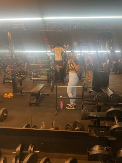 The image depicts a detailed mirror reflection of an active gymnasium in Anápolis, Brazil. The primary subject is a young adult female, positioned in the mid-right, capturing a selfie using a dark smartphone held to her face. She wears a black cropped t-shirt with a yellow graphic, light blue athletic leggings, and black Vans sneakers.

In the reflected background, multiple individuals are present. Directly behind the female, a male is performing pull-ups on a machine, dressed in a light-colored t-shirt and dark shorts. Another male stands further to the right, partially obscured. The gym's interior features dark walls and flooring, likely rubber matting, under bright overhead strip lighting that causes glare on the mirror surface. A white sign affixed to a wall in the upper-left reflection displays Portuguese text: "GUARDE OS PESOS APÓS O USO" (Store the weights after use). A dim red digital display is visible in the upper-middle background.

Fitness equipment is extensively distributed throughout the scene, including various dumbbell racks, barbells on stands, and a black adjustable weight bench positioned in the mid-left. A pair of yellow circular weights or resistance bands are located on the floor to the left of the bench, and a pink water bottle rests on a low rack near the primary subject.
