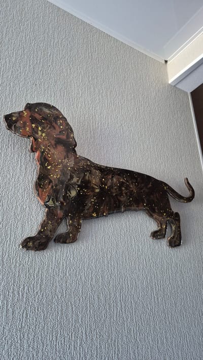 The image shows a dark brown, abstract representation of a cat-like creature with gold flakes embedded in its surface. The creature is depicted in a leaping or running pose, with its body arched and limbs extended. Its tail curls upwards, and its head is turned to the side. The creature's surface has a swirling, marbled pattern, with darker brown and reddish-brown tones intermingling with the gold. The texture of the background is a light gray, textured wallpaper. In the upper left corner, a white door frame and trim are visible, suggesting an indoor setting. The lighting appears to be natural, casting a slight shadow from the creature onto the wall. No text is visible in the image. The overall impression is of a decorative wall art piece, possibly made of resin or a similar material, placed on a textured wall.
