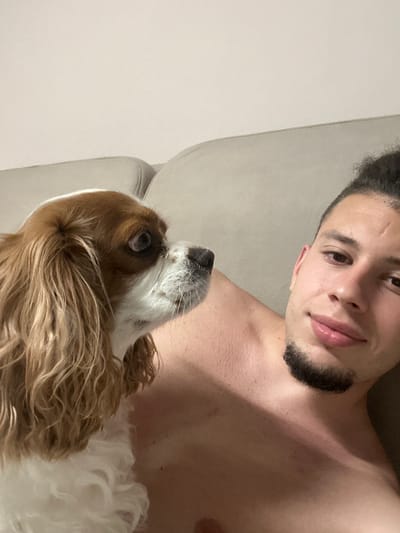 The image displays a young adult male and a Cavalier King Charles Spaniel dog in an indoor setting. The male is situated on the right side of the frame, captured from the head to the upper chest, shirtless, with dark, curly hair pulled back and a goatee. He exhibits a faint smile directed towards the viewer. The dog, a Blenheim-colored Cavalier King Charles Spaniel with long, wavy brown and white fur, occupies the left side of the frame, positioned next to or on the man. Its head is turned to the right, with wide-open eyes looking slightly upwards. The background consists of a plain, light-colored wall and a portion of a light-gray upholstered couch or bed, indicating a domestic environment.