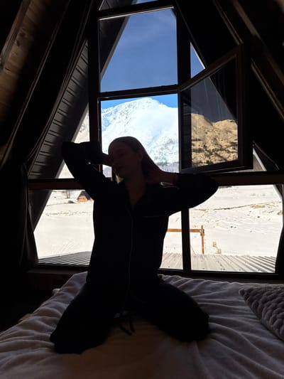 An interior scene in Virak, Montenegro, depicts a female subject kneeling on a white bed, largely silhouetted against a large A-frame window. She wears dark pajamas with light piping and has her arms raised, hands behind her head. The window, constructed with dark wooden frames, has one pane on the right side open inward. Outside the window, a bright, clear blue sky is visible above a prominent snow-covered mountain. Below the mountain, an extensive snow-covered landscape stretches, revealing a small, dark-roofed A-frame cabin in the distant left and a corrugated metal roof structure in the immediate foreground. The interior of the room is dimly lit, with dark wooden paneling visible, while the exterior is brightly illuminated by sunlight. A textured white duvet covers the bed, with a textured pillow partially visible on the right.