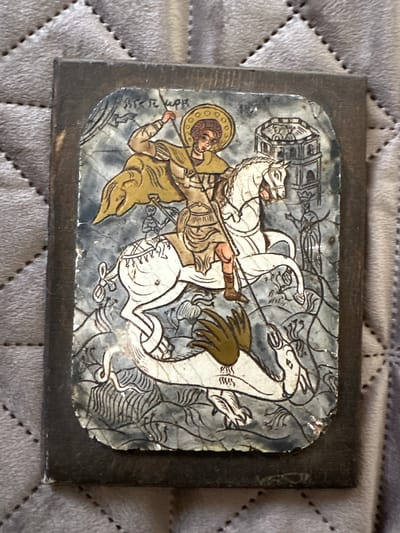 An image shows a religious icon depicting Saint George slaying a dragon. The saint is on horseback, spearing the dragon. In the background, a tower with figures is visible, and a woman with outstretched arms stands to the right. The icon appears to be mounted on a wooden base. There are no discernible landmarks or context to place this image in Marten, Bulgaria.