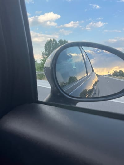 The image shows a view from inside a car, looking out the side window and into the side mirror. The car is traveling on a highway, as indicated by the road markings and the speed blur. The side mirror reflects the scene behind the car, which includes the sky, clouds, trees, and other vehicles on the road. The sky is blue with scattered white clouds, and the sun appears to be setting, casting a warm glow on the clouds. The trees are green and lush, suggesting a rural or suburban setting. There are no people visible in the image. The location is identified as Shtarkovo, Bulgaria.