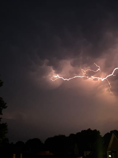 The image depicts a dramatic nighttime scene in Putten, Netherlands, characterized by a powerful lightning strike illuminating the dark, cloudy sky. The lightning, a bright, jagged line of white and orange, cuts horizontally across the middle of the frame, originating from the right and extending towards the left. The clouds are dense and dark, with some showing a rounded, mammatus appearance, suggesting an approaching storm. Below the sky, a silhouette of trees forms a dark, undulating line, representing the natural landscape of the area. In the foreground, the dark shapes of houses and possibly other structures are visible, hinting at a residential or semi-rural setting within Putten. The overall atmosphere is one of intense natural power and the quiet anticipation of a storm.