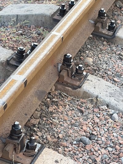 The image shows a close-up view of a railway track, focusing on the fasteners that secure the rail to concrete sleepers. The subjects are the railway components themselves: the steel rail, concrete sleepers, and the metal clips and bolts that hold them together. The scene is a section of a railway line, with ballast stones surrounding the sleepers.
There is no discernible activity or event taking place; the image appears to be a static capture of the infrastructure.
Notable details include the texture of the weathered steel rail, the rough surface of the concrete sleepers, and the black painted bolts and nuts. The ballast is composed of small, multi-colored stones, predominantly reddish-brown and gray. The lighting suggests it is daytime, with no strong shadows indicating a specific time of day. There are no clear location cues beyond it being a railway, other than the provided context of Kant, Kyrgyzstan. No emotions are conveyed.
There is no visible text in the image.