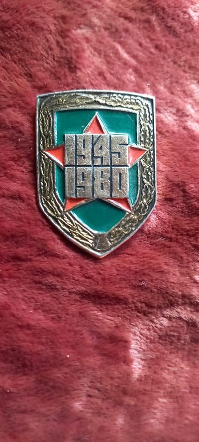 A silver and green shield-shaped medal with red stars and the numbers "1945 1980" in silver is displayed on a red, fuzzy surface, possibly velvet. The medal has a textured silver border with gold-colored detailing. The numbers "1945" are above the number "1980", separated by a horizontal line within the shield. The image is a close-up shot of the medal, with the background blurred. There is no discernible location in Novopavlovka, Kyrgyzstan.