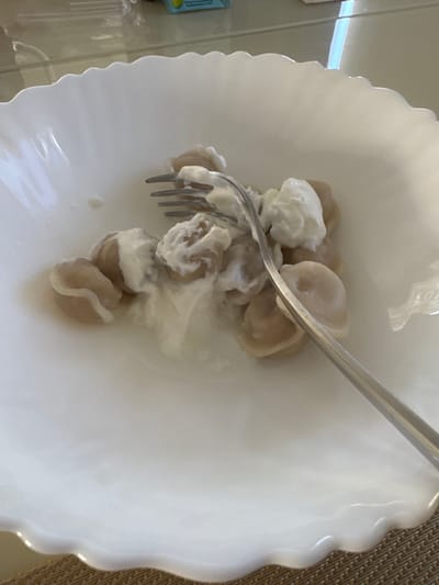 A close-up, eye-level shot shows a white, scalloped bowl filled with pelmeni, a type of dumpling, covered in a generous dollop of sour cream. A silver fork is partially submerged in the food, with its tines picking up some of the dumplings and cream. The bowl sits on a woven placemat. The background is blurred, but appears to be a table surface with reflections. The lighting suggests an indoor setting, possibly a kitchen or dining room, with the natural light filtering in from a window. The image does not provide any specific details or context to locate it in Uyutnoye, Ukraine.